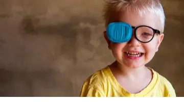 Smiling young boy with blond hair wearing glasses with a blue eye patch covering one eye, commonly used for amblyopia (lazy eye) treatment.