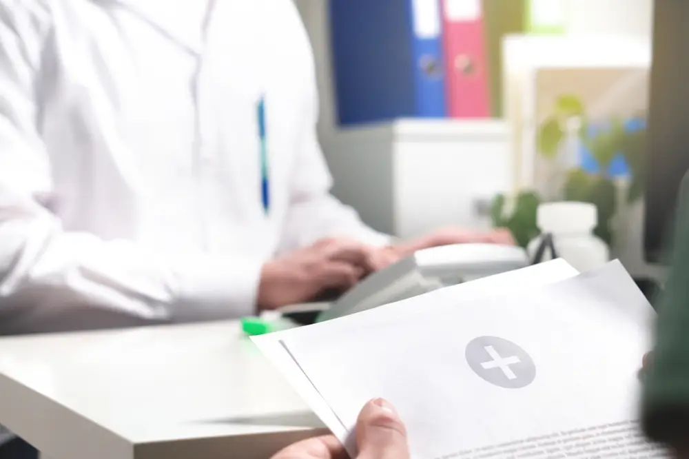 Close up of some medical paperwork held by a patient sitting in front of a desk, behind which sits a man in a white lab-coat.