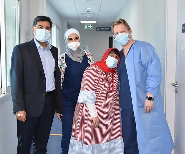 Group of ophthalmologists in Abu Dhabi wearing face masks and standing in a hospital corridor with a smiling patient in a red and white striped dress.